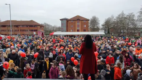 Wai Yee Hong A crowd of people on a winter's day gathered together in front of a stage on which there is a woman in a red coat with her back to the camera