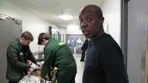 Clive Myrie in a hospital corridor, looking at the camera. Behind him two paramedics are assisting a patient on a trolley