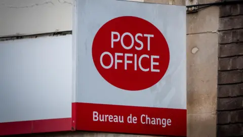 Getty Images An external shop sign of the Post Office. The small, square sign, attached to a light stone wall, is white and has 'Post Office' written in a red circle. Underneath the logo is 'Bureau de Change' written in white against a red rectangular background.