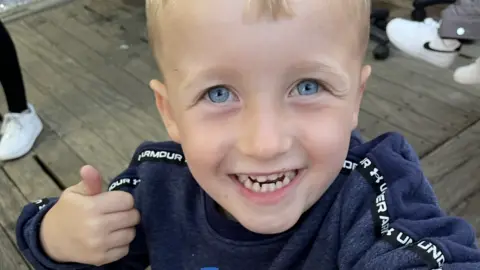 A six-year-old boy, with blue eyes and blonde hair, grins at the camera while giving a thumbs up with his right hand, 