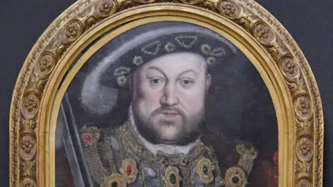 An oil painting of Tudor King Henry VIII with an arched gold frame. The former king is wearing a feathered hat and an ornate gown.