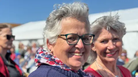 There are two ladies in the picture. One is wearing glasses and smiling at the camera. She has a purple cardigan and a scarf on.
