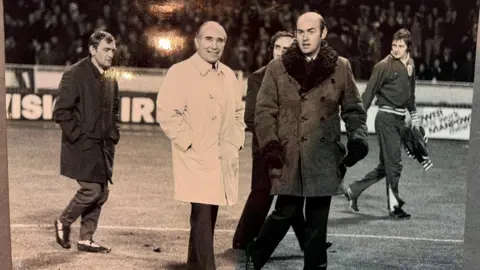 Sir Alf Ramsey and Phillips pictured in long coats walking across a football pitch with players and staff behind them. There are fans in the stands in the background.