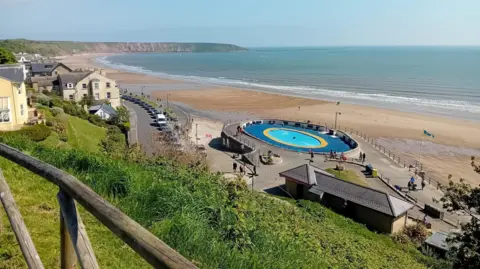 A view of Filey beach and seafront from above. A long sandy beach with a headland in the distance. The tide is out and the sun is shining. Cars and houses line the promenade, with a large paddling pool situated on the right, overlooking the beach.