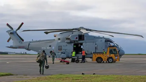 Crown Copyright Crew in yellow and pink high vis are seen around a helicopter which has its doors open and a yellow truck is parked to the right. A person in miliary green green is walking away from he camera carrying a backpack and some equipment in their other hand.