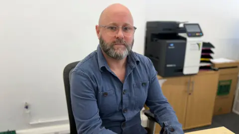Bald man with grey facial hair and glasses blue shirt sat at desk