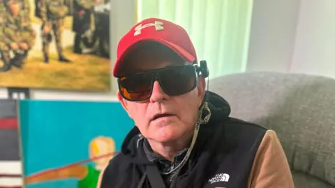 a man wearing dark sunglasses, a red hat and black gillet sitting on a couch