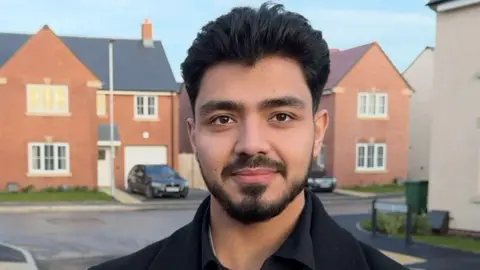 A man with short black hair, a beard and wearing a black shirt, jacket and lanyard, stands in the middle of a housing estate.