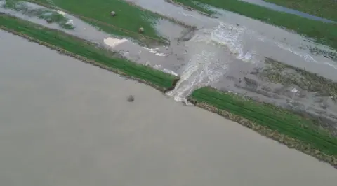 ovoto.co.uk An overhead picture of the breach in the River Welland at Crowland. Water can be seen gushing through a gap that has emerged in the riverbank and into the field beyond. 