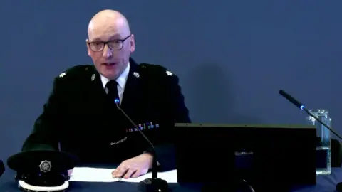 Omagh Bombing Inquiry Norman Haslett at the Omagh Bombing Inquiry. He is wearing a police uniform and sitting in front of a computer and microphones. He is bald and wears glasses.