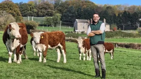 Wye Organic A man in green trousers, a blue shirt and a dark green bodywarmer stands in a grassy field. He is surrounded by brown and white Hereford cattle.