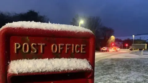 BBC A red post box, with the words post office in gold writing, has snow sitting on top of it. It is next to a snow-covered road.