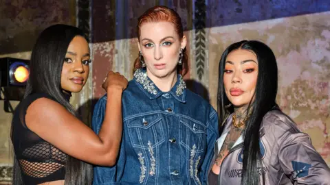 BBC Studios Sugababes, left to right, Keisha in a black mesh top, with her right hand on the shoulder of Siobhan, centre, wearing a blue denim jacket, with Mutya to her right with a shiny grey sports jacket.