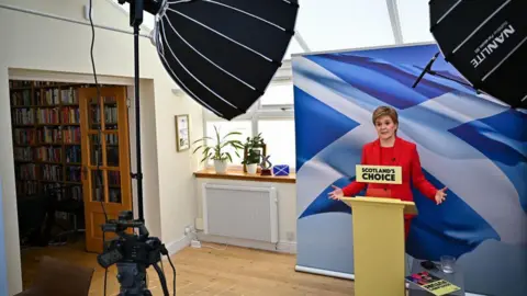 Getty Images Nicola Sturgeon standing at a podium in her conservatory with a large saltire in the background. She is wearing a red trouser suit.