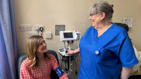 Patient, Hannah Robinson, wearing a red and white checked dress sitting in a hospital room smiling at a nurse standing in front of her, wearing a blue tunic and taking her blood pressure