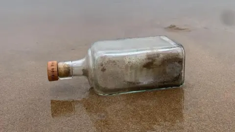 Mike Nicholls A bottle with a cork. There are ashes inside the bottle and a note. It is lying on its side in the sand. 