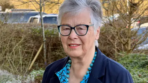 JAMIE NIBLOCK/BBC Janet Beacham stands outside her office in Colchester. She is in front of a hedgerow and a few trees without leaves. There are cars parked behind that. She is wearing a navy blue jacket and has a light blue mottled shirt and matching blue beads around her neck.