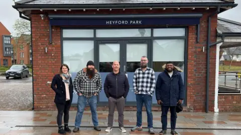 Heyford Park Community Action Group Five people are standing in from of a stone building, with the words Heyford Park aboue them. A woman stands to the left, then four men. It looks like it has been raining.