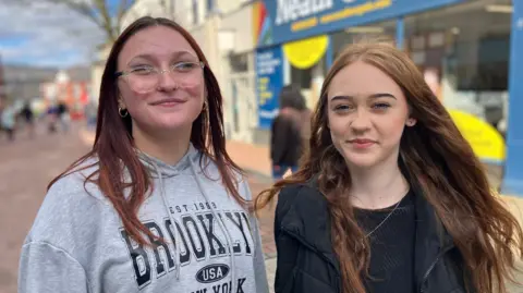 A girl with long died red hair and glasses, wearing a grey hoody with Brooklyn written on it in black letters and smiling at the camera. Next to her is another girl with long reddish-brown hair and nose piercings, wearing a black jumper and black jacket and smiling at the camera. Behind them is a shopping street.