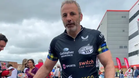 Image of a man in cycling gear. In the background can be seen a crowd of people and the outside of a large stadium building