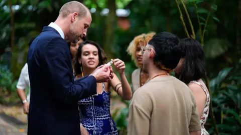 PA Media The Prince of Wales holds a small bottle he's been given as he meets with young Brazilian leaders who surround him in a semi-circle. William is facing them, head bent to look closely at the bottle. He is in profile to the camera. A young woman has her finger on the top of the bottle and is speaking to the prince. 