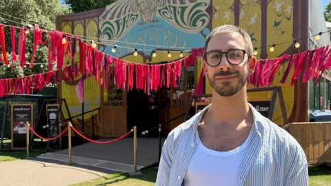 James McConnell/BBC Jon smiles at the camera on a sunny day, surrounded by the pink and yellow festival decorations. He has short brown hair and wears glasses, and has dressed in a white vest with a striped shirt open over the top.