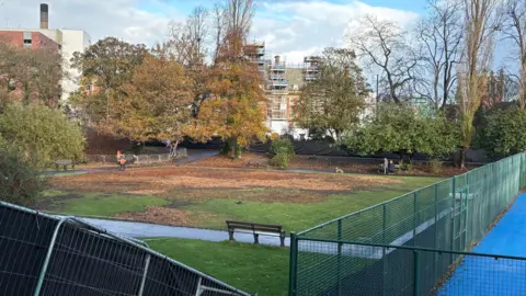 Local Democracy Reporting Service The section of the park where the maze previously stood. The blue tennis and basketball courts are partially visible in the right hand side of the image and are surrounded by a tall, green fence. The area where the maze was now consists of low cut grass with large brown patches. There are paths and trees around the area.