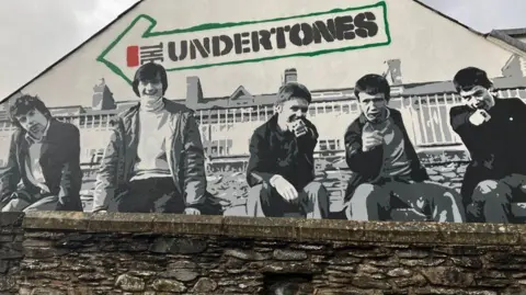 A black and white mural of five men, dressed in 1970s gear. They are sitting on a bench. The two to the left of the picture are looking into the camera. The other three are pointing at the camera.