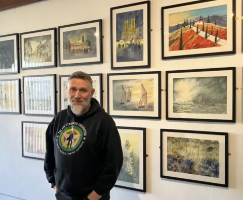 Lara King Tim has short dark hair and a grey beard. He is wearing  black hooded top with a green circular print and is standing in front of a wall on which a series of framed paintings are hanging.