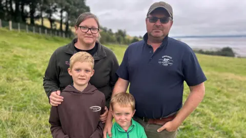 BBC A family - a man, woman and two young blonde boys - standing together in a field, all looking into the camera. The parents have their hands on the boys' shoulders. They are all dressed casually and in warm clothing, apart from the man who is wearing a cap, sunglasses and a blue polo shirt. He has one hand in his trouser pocket.