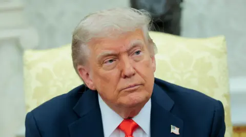 Donald Trump is looking away from the camera. He's wearing a navy suit with a white shirt and a red tie. He's sitting in a yellow chair.