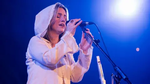 A woman with brown hair is wearing a white linen long-sleeved top with the hood up, and is singing into a black microphone which is on a stand. The background is dark blue on stage