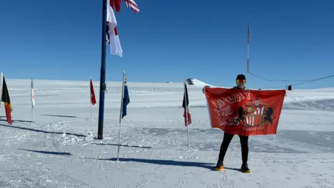 Paul Holborn standing in Antarctica holding a big, red Sunderland AFC flag. He's wearing colourful sunglasses and a cap. The ground and horizon is covered in snow. Various flags stand around him.