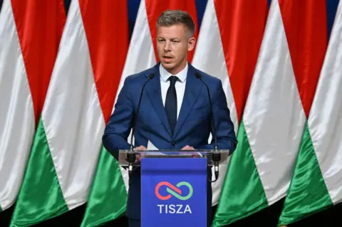 Peter Magyar holds a press conference in Budapest. He wears a blue suit and stands against a backdrop of the Hungarian flag.