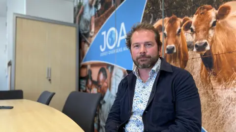 Ed Lewis is sitting at a conference table in an office setting. The background features a large wall mural with images of cows and people, along with the text 'JOA' and 'Jersey Overseas Aid.'