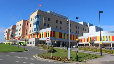 UHNM A hospital with ambulances parked outside it. The hospital building is several storeys high, and has red and yellow cladding on the lower part.