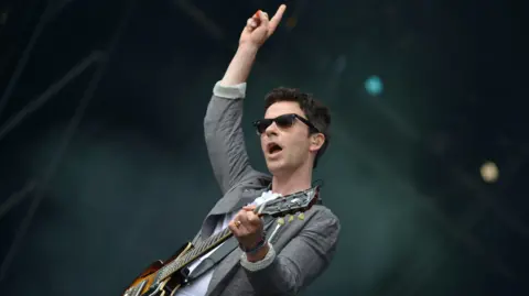 Kelly Jones of Stereophonics on stage. He has short dark hair, wears sunglasses, and is wearing a white t-shirt and grey blazer. He is holding a guitar and his hand is a aloft, pointing to the sky.