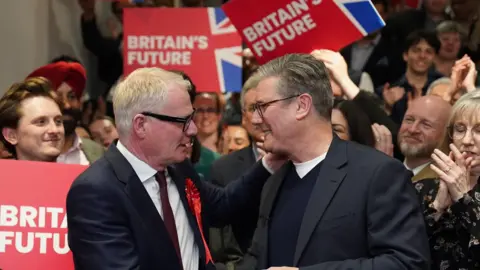 Sir Kier Starmer and Richard Parker, both wearing suits, shake hands and look towards each other amidst a crowd of clapping people. Some of them are holding signs that say Britain's future