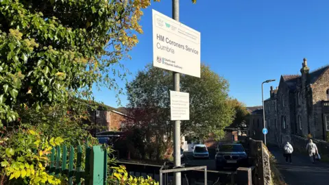 BBC A general view of the entrance to the coroner's office in Cockermouth. There is a sign that says 'HM Coroners Service Cumbria' outside the entrance to a car park that leads to a low building.