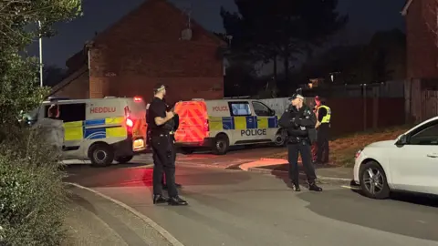 BBC Three police officers can be seen standing on the street, with two police vans behind them. It is dark, and one van has its brake lights on. Two police officers are wearing a black uniform with black caps on. A third is slightly in the background facing away, wearing a yellow fluorescent vest. 