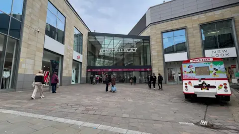 The exterior of a shopping centre with an ice cream van on the pavement on the right-hand side.