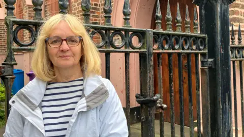 Leanne Harper/BBC A middle aged woman with blonde hair, glasses and wearing a stripey blue and white top under a white coat, smiles at the camera outside a set of black iron gates in front of a church building. 
