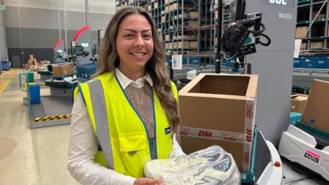 Ben Schofield/BBC Diana Kovacs looking down the camera, standing inside a warehouse. She is wearing a yellow, high-visibility gilet over a white shirt and light brown knitted tank top. She is smiling, showing her teeth. Her long, wavy brown hair falls over one of her shoulders on the right of the image. She is holding a pair of trainers, wrapped in a cellophane bag. Next to her is a cardboard box, at about chest height. The box is sitting on top of a small robot, with another robot queuing up behind, also carrying a box. Further in the background are other workstations and shelving holding other boxes.
