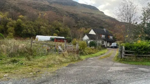 A house in Dornie, Ross-shire. There is a gravel path with some grass in the centre leading up to the house, which is white with a dark roof and two windows facing out the front surrounded by light brown trim. A dark-coloured van is parked outside. The house is surrounded by greenery and a bare tree. In the background is a hill under a cloudy sky.