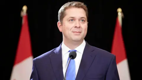 Andrew Scheer is photographed speaking at a podium. He is wearing a navy blue suit and a light blue tie. Behind him are two Canadian flags. He has short light brown hair and blue eyes.