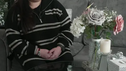 BBC A woman with her face out of frame sits on a grey sofa with her hands clasped together in a black and white jumper. She is sat in front of a vase of flowers, a candle and a box of tissues set on a table.