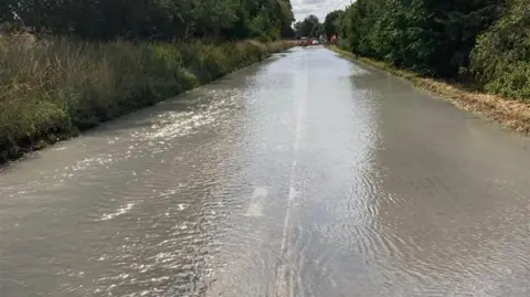 A road pictured from the middle of the carriageway, disappearing into the distance. It is completely underwater and you can barely make out the white lines. It is hedged by verges and trees on both sides and in the distance is a series of vehicles. 
