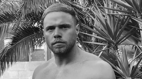 Family photo Corey pictured in black and white. He is bare chested and standing in front of a wall and a palm tree.