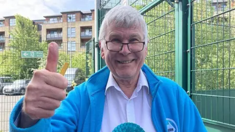A man with grey hair and glasses, wearing a pale shirt under a blue fleece, gives the camera a thumbs-up.