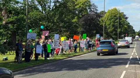 A busy road with protesters with placards and signs lining the grass verge.
